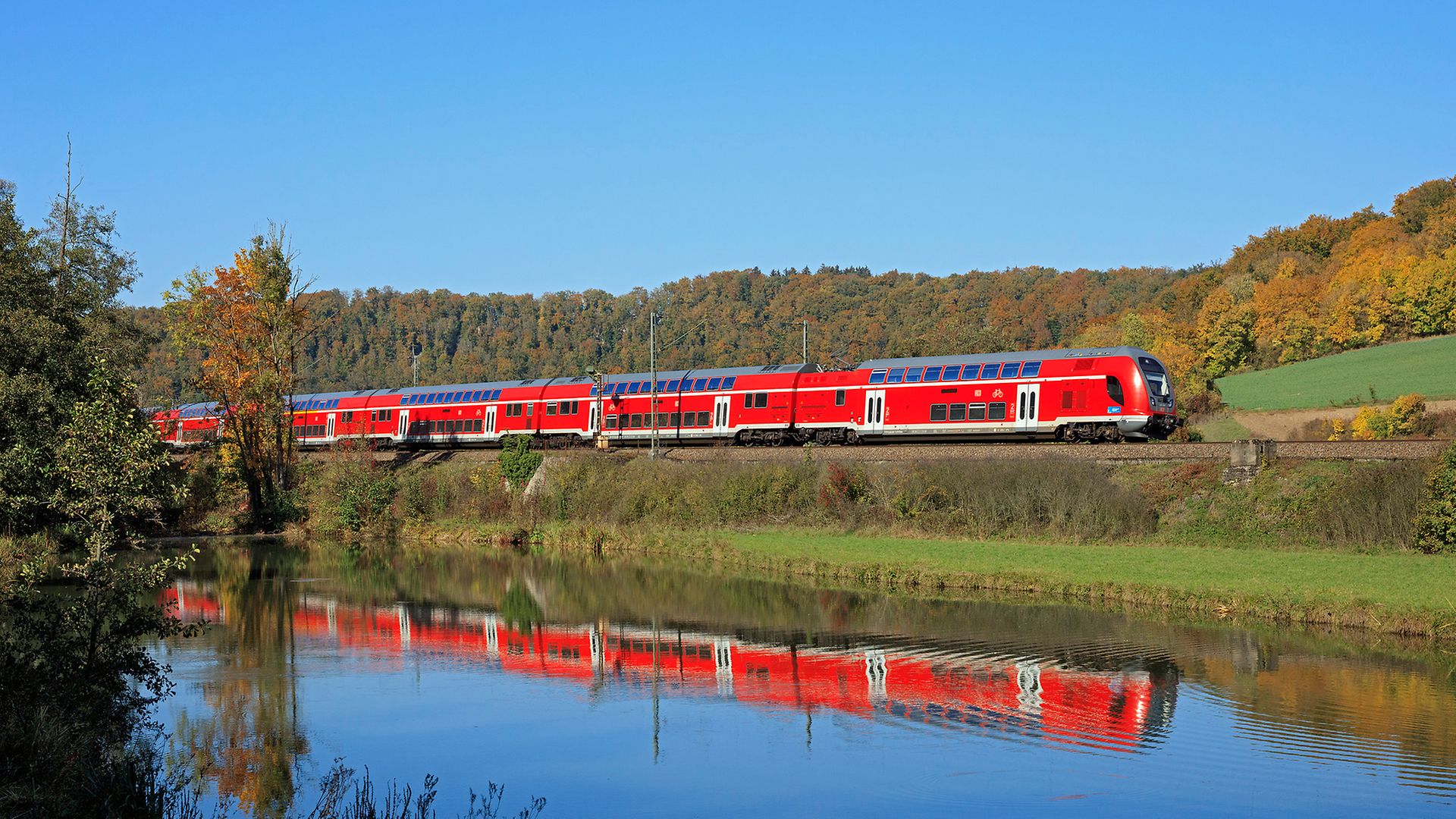 Roter Zug fährt durch grüne Landschaft neben einem See in dem sich der Zug spiegelt