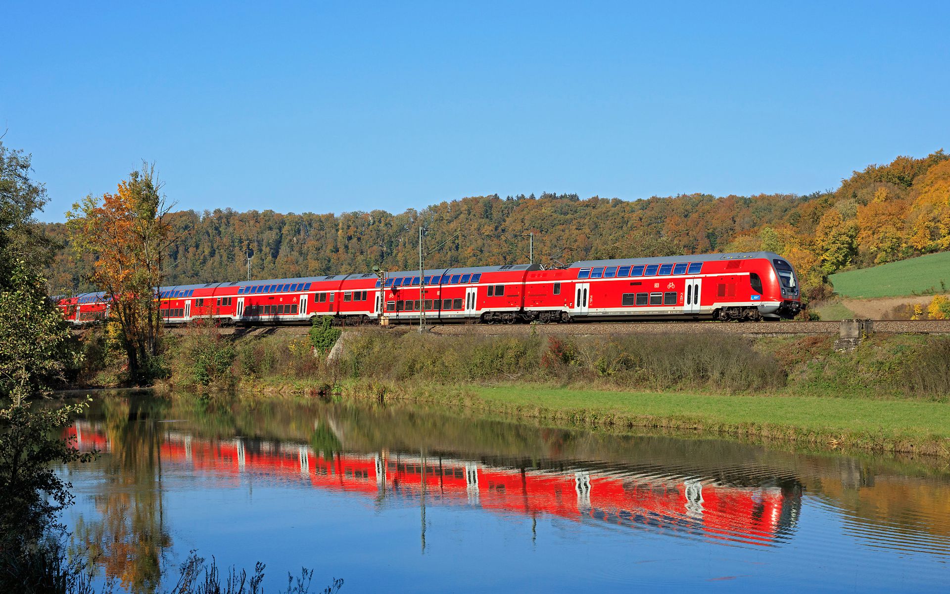 A red train travels through green countryside alongside a lake in which the train is reflected