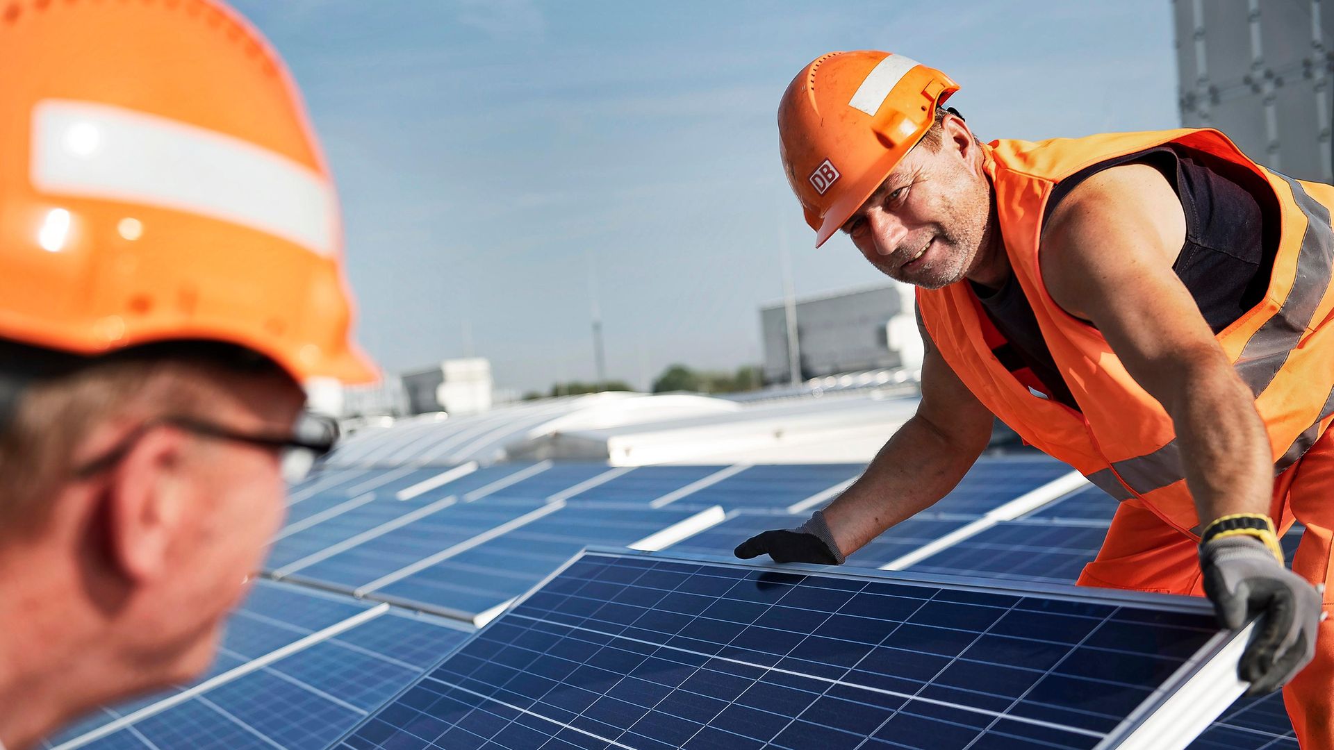 Zwei Mitarbeiter in Schutzkleidung verlegen Photovoltaik-Platten auf einem Dach bei blauem Himmel. 