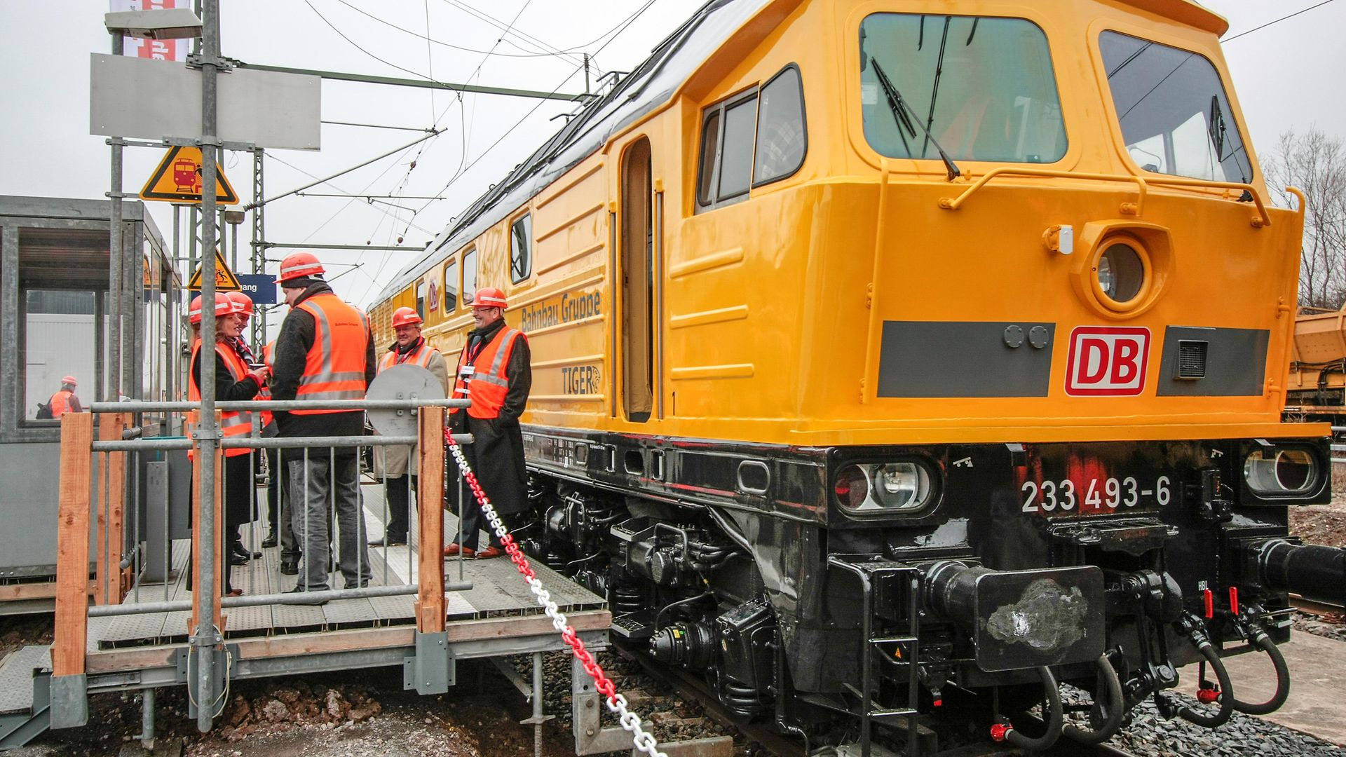 Gelbe Maschine der Baureihe 233 neben einem Haltepunkt an dem Mitarbeiter stehen