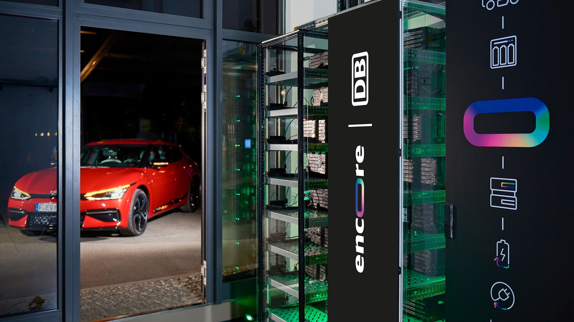 Indoor Batteriespeicher mit Schriftzug "encore | DB", hinter einer geöffneten Glastür roter KIA PKW